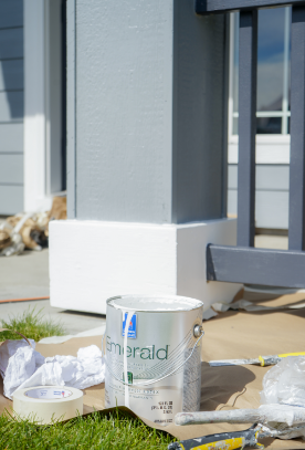 A paint can labeled "Emerald" and painting supplies are scattered on the ground near a painted porch post. The area is covered with protective paper to prevent spills. The exterior porch is painted gray and white, and the weather appears sunny with clear skies in the background.