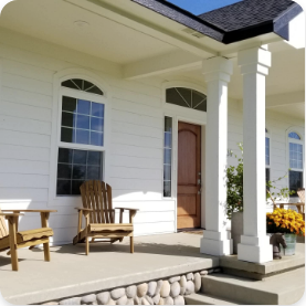 A quaint porch of a white house with a wooden front door, two large windows, and an overhang supported by two columns. The porch features two wooden chairs, a small side table, and potted flowers—all bathed in sunlight. Ideal for cozy moments or perfect as inspiration for commercial painting services.