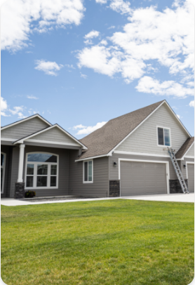 A modern, single-story gray house with a neatly trimmed lawn under a bright blue sky with scattered clouds. The house, showcasing expert exterior painting services, has a large window and a double garage with a ladder leaning against its side. White trim details accentuate the home's design.