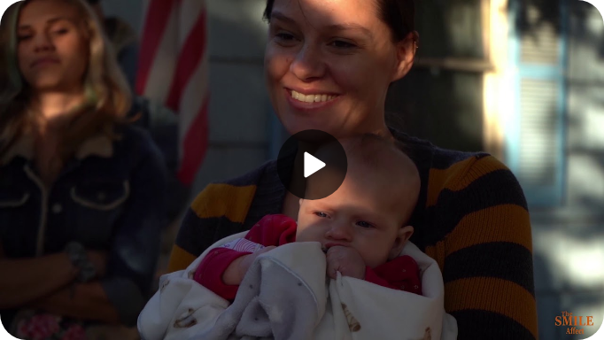 A woman smiles as she holds a baby bundled in a blanket, enjoying the natural light of the beautiful exterior. Another person stands in the background with an American flag visible beside them, highlighting a picturesque outdoor scene.