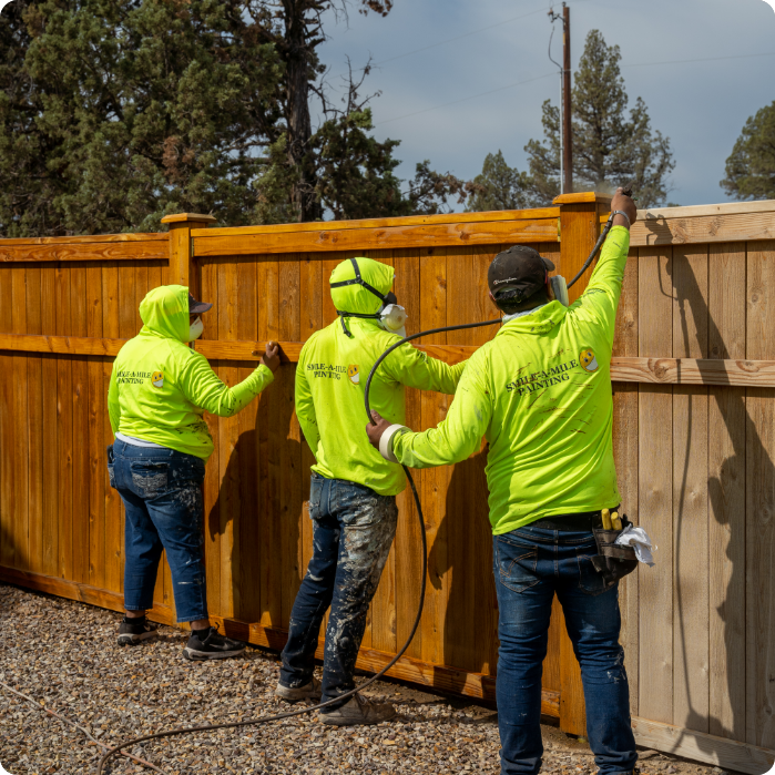 Three workers wearing bright green shirts with logos are providing commercial painting services outdoors. They each hold painting equipment and are applying a brown finish to a wooden fence. The area, surrounded by trees, appears to be in a semi-rural location.