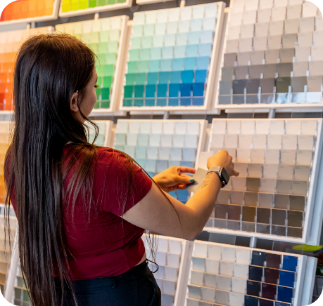 A person with long hair wearing a red shirt and black pants is selecting color swatches from a display wall filled with various shades arranged in rows. The wall includes a wide range of colors, perfect for anyone seeking inspiration for commercial painting services, covering both interior and exterior designs.