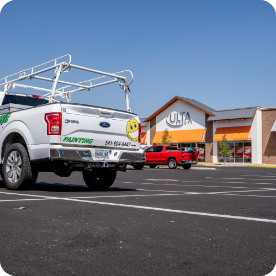 A white truck with a painting company’s logo and contact number is parked in a nearly empty strip mall parking lot, ready to offer exterior and interior commercial painting services. The truck has a ladder rack on top. In the background, there is an Ulta Beauty store and a red truck. The sky is clear and blue.
