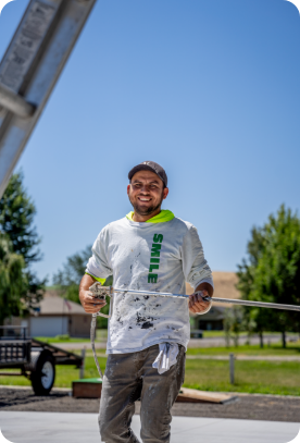 A man in a white, paint-splattered shirt and cap is smiling while holding a measuring tape outdoors. The sky is clear and blue, and there are trees and a house in the background. He appears to be offering exterior or interior painting services at a construction site.