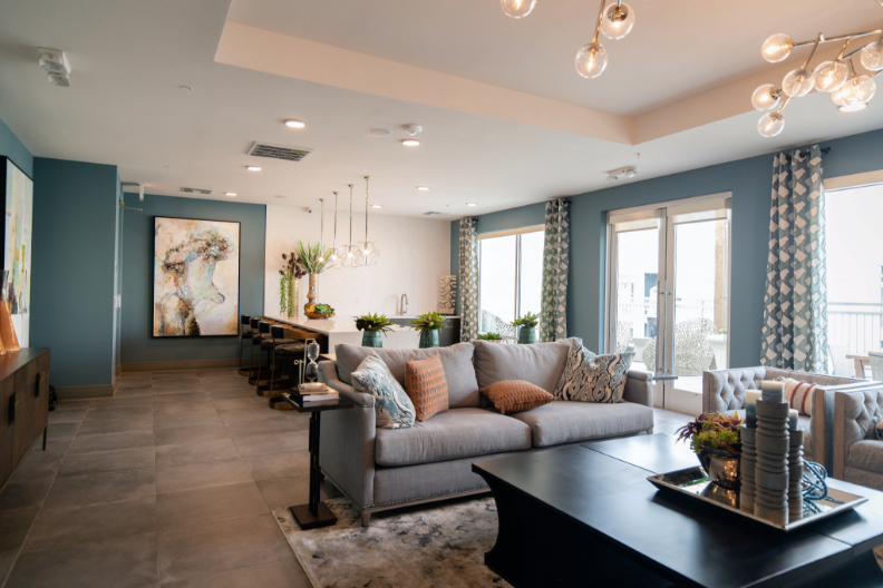 A modern living room with blue walls and large windows. It features a gray sofa with orange and patterned throw pillows, a dark wooden coffee table with books and decor, and a kitchen area with a long island and bar stools. Contemporary light fixtures hang from the ceiling, showcasing top-tier commercial painting services.