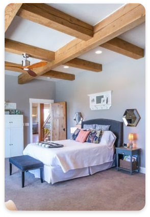 A cozy bedroom features a wooden bed with white linens and colorful pillows. Exposed wooden beams highlight the ceiling, adding rustic charm. Nightstands with lamps flank the bed, and a bench sits at its foot. Hexagonal mirrors and a small shelf adorn the walls, embracing both exterior and interior design elements.
