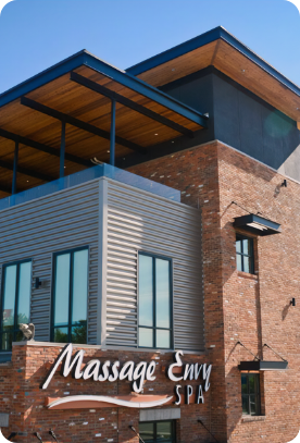 A modern two-story building with a mixed façade of brick and metal siding. The building, offering commercial painting services, features a sign reading "Massage Envy SPA" in white lettering on the lower section. The roof has an overhang and the upper story has large windows. The sky is clear and blue.