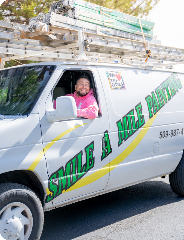 A man in a pink hoodie is smiling while sitting in the driver's seat of a white van loaded with ladders on the roof. The van, offering exterior and interior commercial painting services, has "Smile A Mile Painting" written on its side and a sign that reads "We ♥ Tri-Cities". The background features trees.