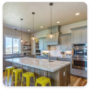 A modern kitchen with grey cabinets, stainless steel appliances, and a large island with a white countertop. The island has four yellow bar stools underneath. Three pendant lights hang above the island, and the kitchen is well-lit with natural light from a window, perfect for entertaining or gathering after commercial painting services.