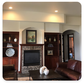 A cozy living room with built-in wooden shelves on either side of a stone fireplace. There are various decorative items on the shelves, a commercial painting above the fireplace, and dark brown leather couches around a coffee table in the center.