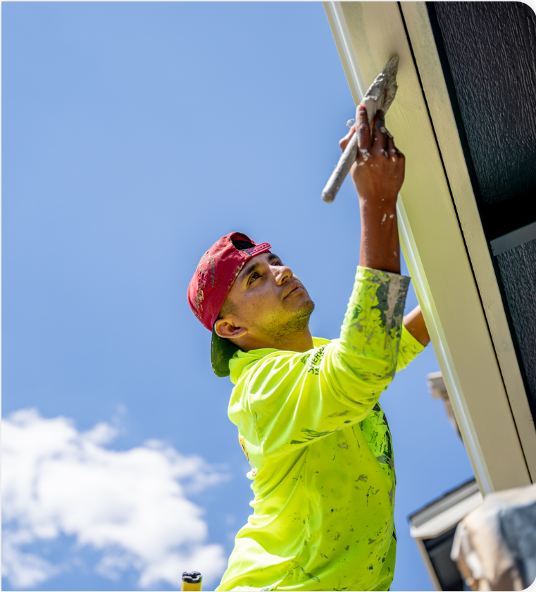 A worker wearing a red cap, neon green shirt, and gloves is applying paint with a brush to the house’s exterior trim while standing on a ladder against a clear blue sky, showcasing expert commercial painting services.