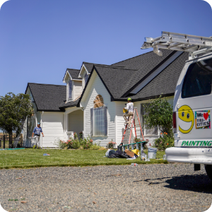 Workers painting the exterior of a white house with a black roof. One worker stands on a ladder, another on the ground. A white van with a smiley face logo and "We Love Our Cities" sticker is parked nearby. Lawn equipment and paint cans are scattered in the yard, showcasing their commercial painting services.