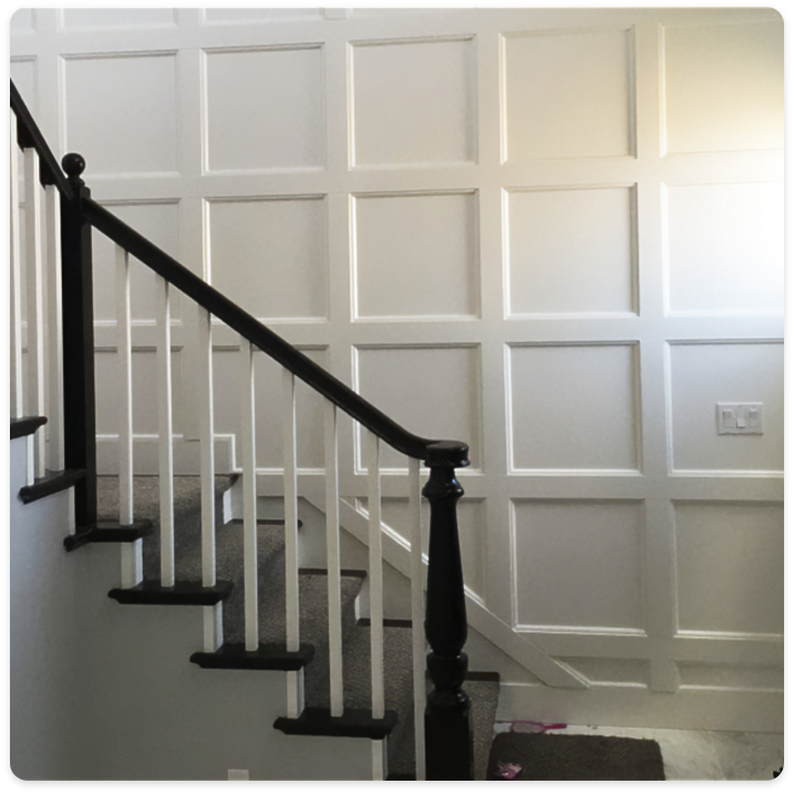 A staircase with dark wooden handrails and white spindles steps up against a wall adorned with white wainscoting in a square paneled pattern. A small section of a carpet is visible at the bottom, along with a light switch on the wall, highlighting the crisp finish provided by our interior painting services.