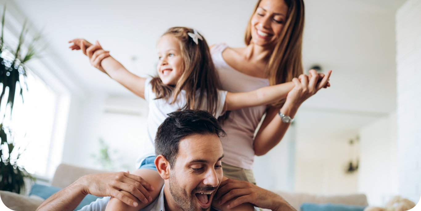 A man, woman, and child are shown in a joyful moment at home. The man, in the foreground, has the child sitting on his shoulders while the woman stands behind them with a smile, holding the child's hands. The bright room filled with natural light highlights their happiness and showcases stunning interior painting services.