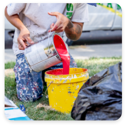 A person wearing paint-splattered clothing pours red paint from a can into a yellow bucket. They are outdoors near a vehicle, likely setting up for some commercial painting services. The ground around them has patches of grass, and a black plastic bag is visible in the foreground.