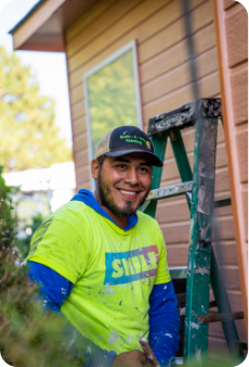 A man wearing a yellow shirt and a black cap smiles at the camera while sitting on a step ladder. He appears to be involved in painting, with visible paint marks on his clothes and hands. Behind him is the exterior wall of a house and some greenery, showcasing commercial painting services in progress.