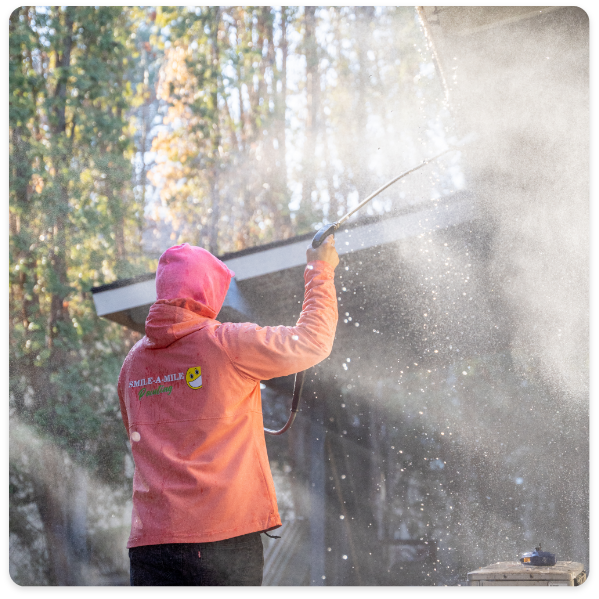 A person in a pink hoodie uses a pressure washer outside with sunlight streaming through surrounding trees. There is mist and spray visible, creating a hazy effect, almost as if prepping the exterior for commercial painting services. The hoodie displays a smiley face logo and text that is not fully readable.