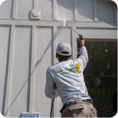 A person wearing a long-sleeve shirt and cap is power washing the exterior wall of a building. They are holding a power washer aimed at the siding, with water spraying out to clean the surface in preparation for commercial painting services.