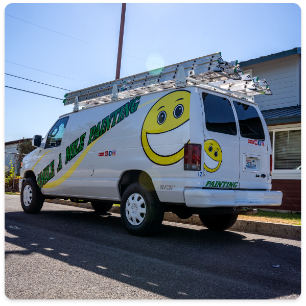 A white van with large yellow smiley faces painted on its sides and rear, along with the text "Smile A Mile Painting," offers both exterior and interior painting services. Two ladders are secured on the roof of the van. The vehicle is parked on a residential street near a house.