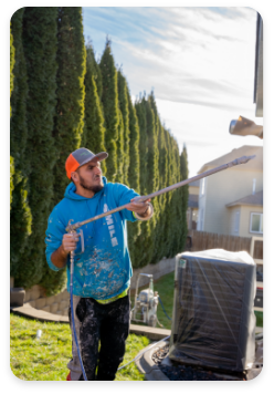 A man in a blue hoodie and orange cap is painting the exterior of a house. He stands on a lawn, holding a long paint roller extended upward to reach the house wall. There are tall conifer trees and a covered outdoor unit behind him, exemplifying professional commercial painting services at work.