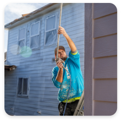 A person in a blue hoodie and cap is standing on a ladder, painting the exterior of a house with a paint sprayer. The large windows are partially covered with protective plastic sheets, showcasing the meticulous approach typical of professional commercial painting services.