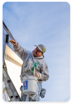 A painter wearing a cap and paint-splattered clothes stands on a metal ladder while providing commercial painting services to the exterior trim of a building. He holds a paint roller in one hand and a paint bucket in the other. The clear blue sky is visible in the background.