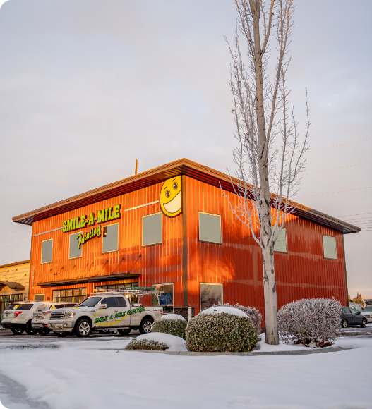 A bright orange building with a large, smiling face sign and the text "Smile-A-Mile" on the exterior. Several vehicles are parked in front, likely for their popular commercial painting services. Leafless trees and snow-covered ground surround the building. The sky is overcast.