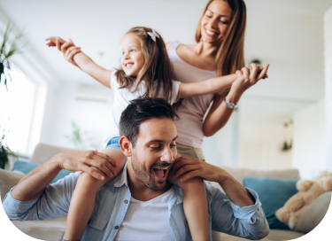 A young girl sits on her father's shoulders as he smiles and holds her legs. Her mother stands behind them, holding the girl's arms out wide. The family appears happy and playful in a well-lit, modern living room, freshly painted by top-notch residential painting service company that bring warmth to their home.