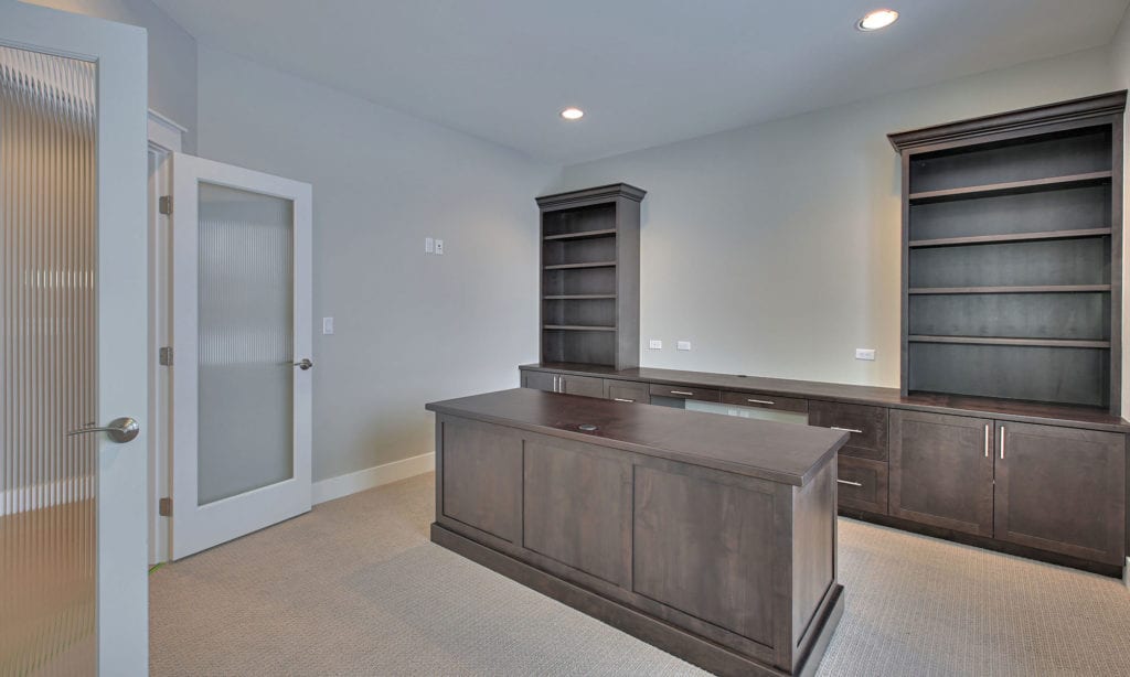 A modern, unfurnished office space featuring a wooden desk in the center. On either side of the desk are built-in dark wood bookshelves and cabinets against light-colored walls. The door, partially open, has frosted glass panels. The floor is carpeted, ideal for interior commercial painting services.