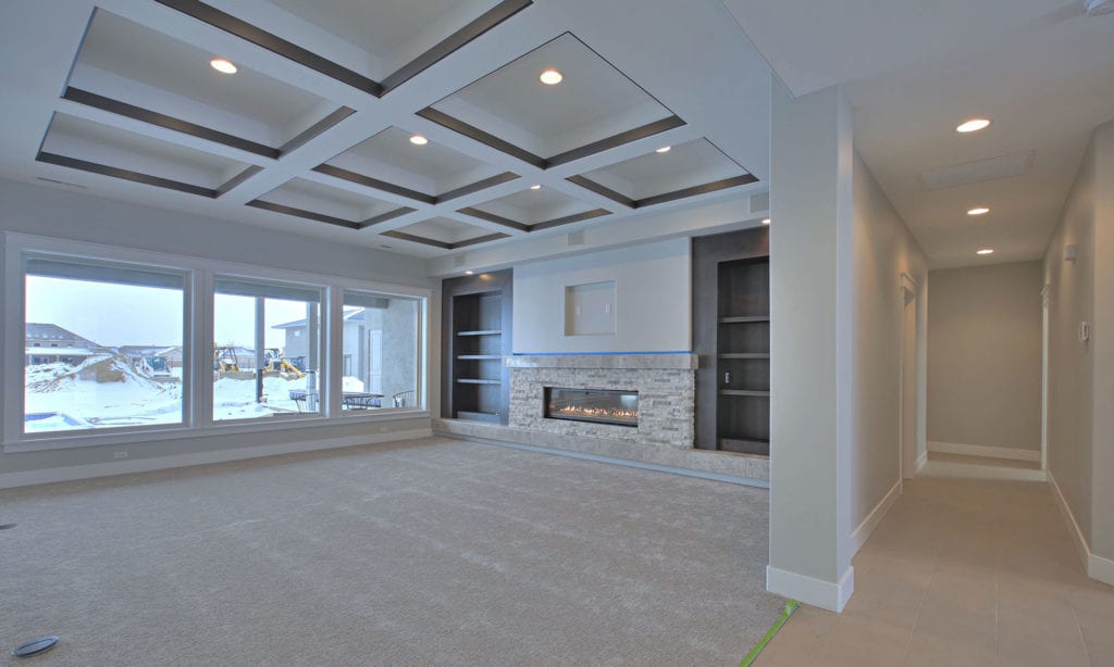 A modern living room with beige carpet flooring and a recessed ceiling with geometric patterns. Large windows provide natural light and a view of a snowy landscape. The focal point is a sleek fireplace embedded in a wall with built-in shelves on either side, showcasing impeccable interior design.